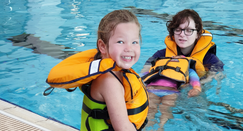a young girl in a life jacket sitting on the side of the pool and an instructor and second girl in the water floating
