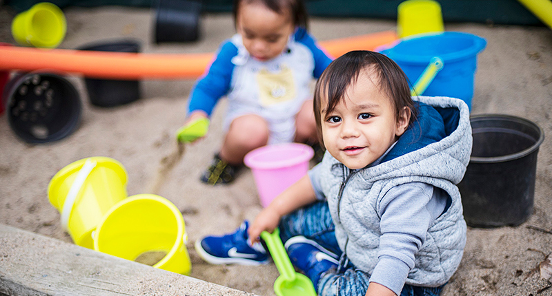 A little boy wearing a grey hoodie plays in the sandpit at Kauri Kids.