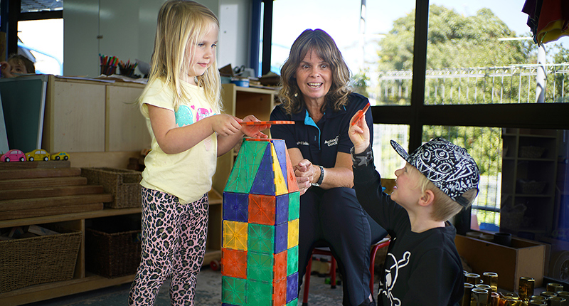 A caregiver wearing a council uniform supervises while two children play with colourful building blocks.