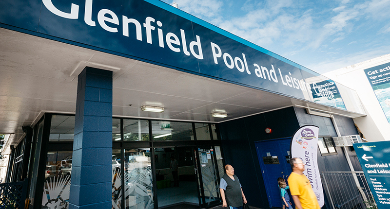 Closeup angled shot of Glenfield Pool and Leisure Centre showing three customers leaving the building, the front sliding doors, blue brick pillar and main signage