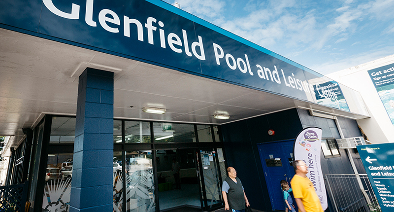 Closeup angled shot of Glenfield Pool and Leisure Centre showing three customers leaving the building, the front sliding doors, blue brick pillar and main signage