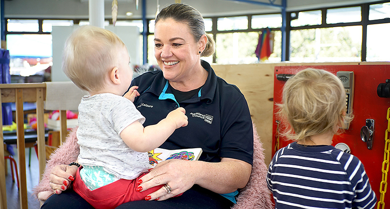 A caregiver wearing a council uniform sits on a pink fluffy beanbag with a child on her lap while another child stands with his back to the camera at her side.