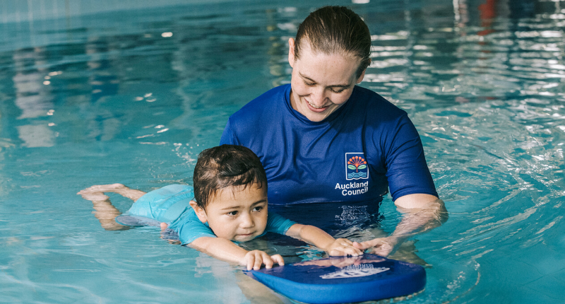 A female Learn to Swim instructor holds a kickboard while she teaches a young child how to swim in the pool