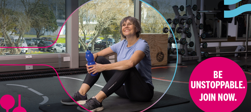 Woman sitting on a gym floor, holding a drink bottle and smiling.
